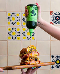 Closeup of hands holding a sandwich on a cutting board while pouring graza onto the sandwich in front of decorative kitchen tile